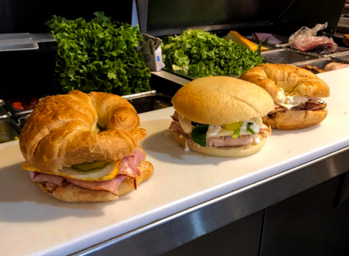 Three sandwiches on a counter: a croissant with ham and pickles, a turkey sandwich with lettuce, and a croissant with roast beef.