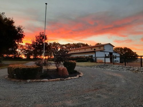 A scenic sunset over a gravel driveway leading to a white building, surrounded by trees and landscaped bushes.