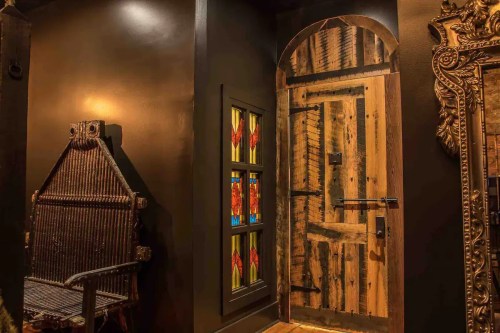 A rustic wooden door with a decorative stained glass window, next to an ornate chair in a dimly lit room.