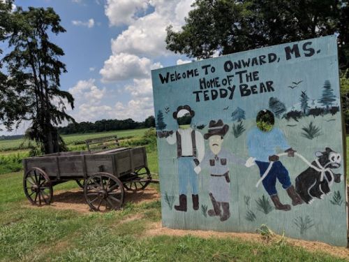 Welcome sign for Onward, MS, featuring painted figures and a wooden wagon against a sunny, rural backdrop.