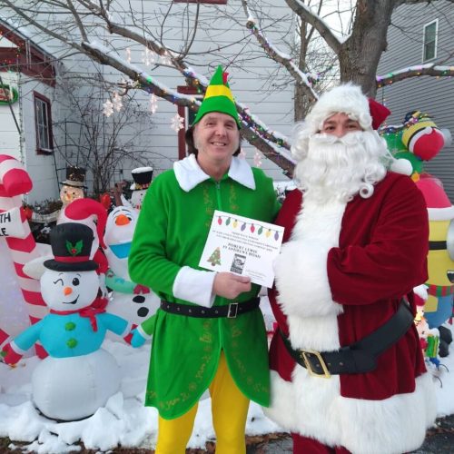 A man in an elf costume and Santa Claus hold a festive certificate in front of colorful holiday decorations.