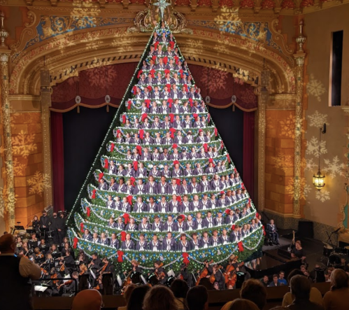 A festive Christmas tree made of performers in a theater, with an orchestra in front and snowflakes in the background.