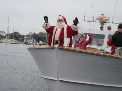 Santa Claus waves from the bow of a boat, accompanied by a child in a winter hat, with a harbor in the background.