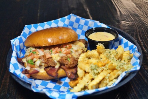 A sandwich with fried chicken, cheese, and toppings, served with crinkle-cut fries and a side of dipping sauce.
