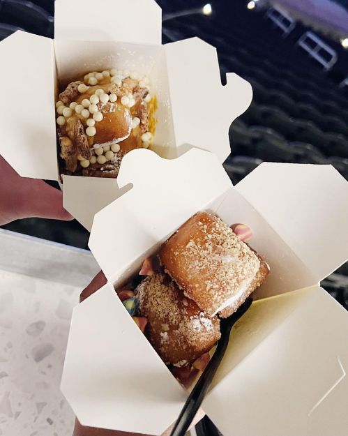 Two takeout boxes held in hands, each containing dessert treats topped with colorful sprinkles and powdered sugar.
