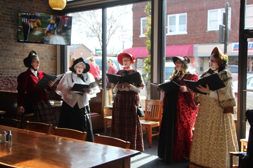 A group of five women in vintage dresses singing indoors, with a TV showing a sports game in the background.