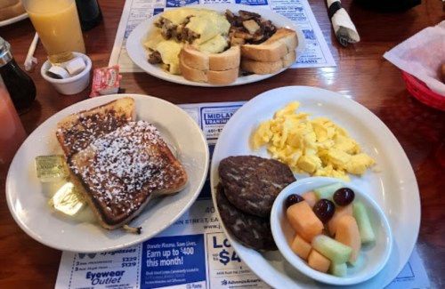 A breakfast spread featuring French toast, scrambled eggs, sausage patties, and fruit on a wooden table.