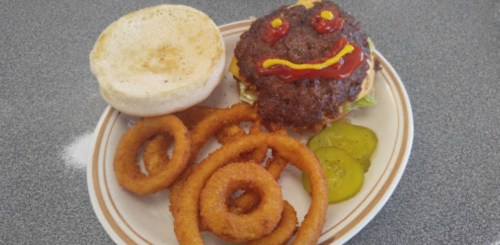 A burger with a smiley face made of ketchup, served with onion rings and pickles on a plate.