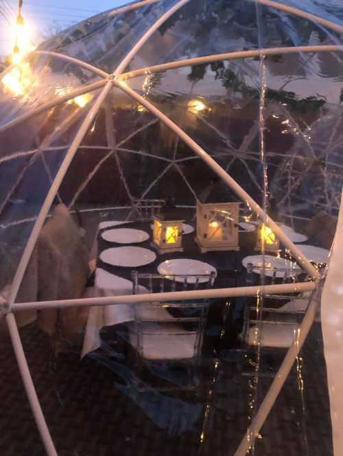 A cozy dining setup inside a transparent dome, featuring a table with lanterns and elegant tableware.