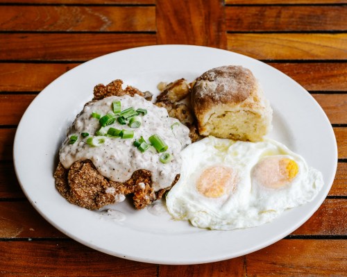 A plate of fried chicken topped with gravy, two sunny-side-up eggs, and a biscuit, served on a wooden table.