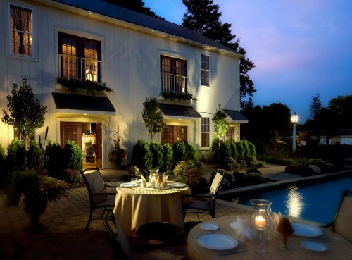 A beautifully lit outdoor dining area by a pool, with a charming house and lush greenery in the evening twilight.