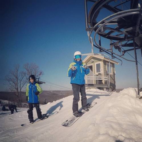 Two children in winter gear stand on a snowy slope, preparing to ski, with a ski lift in the background.