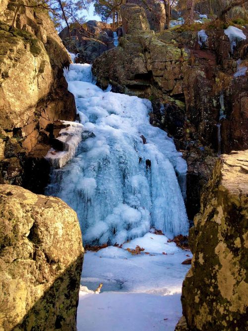 A frozen waterfall surrounded by rocky cliffs and snow, with icicles hanging from the ice-covered surface.