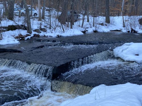 A serene winter scene of a flowing stream with a small waterfall, surrounded by snow-covered ground and trees.