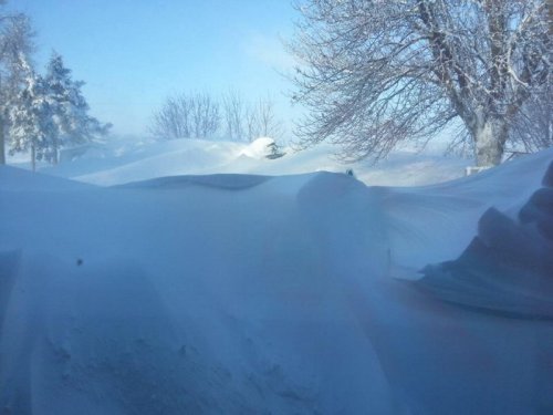 Snow-covered landscape viewed through a window, with drifts obscuring the view and a bright blue sky above.