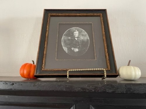 A framed black-and-white portrait on a shelf, flanked by a small orange pumpkin and a white pumpkin.