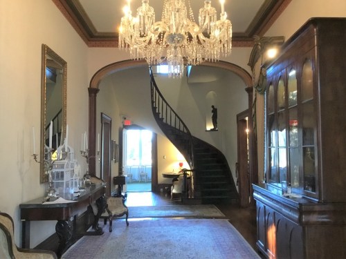 Elegant hallway featuring a grand staircase, chandelier, and antique furniture, with light streaming in from a doorway.