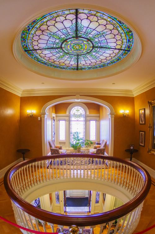 A grand foyer with a stained glass ceiling, elegant seating, and a circular staircase, featuring warm, golden walls.