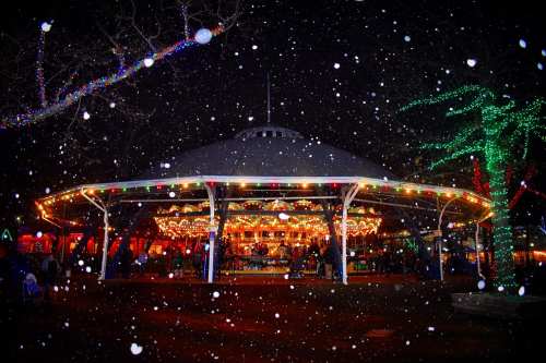 A festive carousel illuminated with lights, surrounded by falling snow and decorated trees at night.