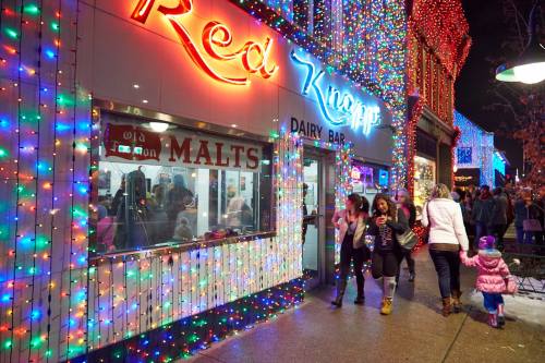 A festive street scene with colorful holiday lights, featuring a dairy bar and people walking by.