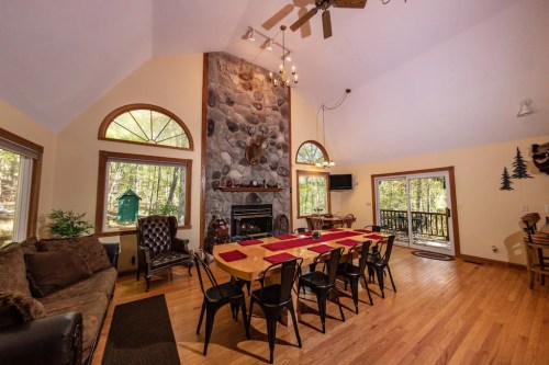 Cozy dining area with a large table, stone fireplace, and large windows overlooking a forested view.