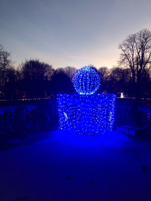 A large blue illuminated gift box with a spherical decoration on top, set against a twilight sky and trees.
