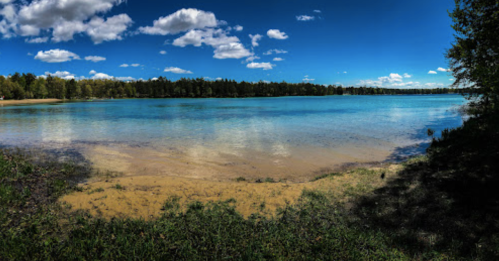 A serene lake surrounded by trees under a bright blue sky with fluffy clouds, reflecting on the water's surface.