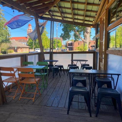 Outdoor seating area with colorful chairs and tables under a wooden canopy, surrounded by trees and buildings.