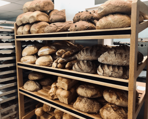 A wooden shelf filled with various types of freshly baked bread, including loaves and rolls, in a bakery setting.