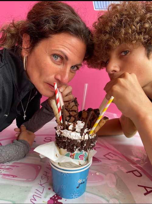 A woman and a boy share a large, decorated ice cream sundae with colorful straws, smiling at the camera.