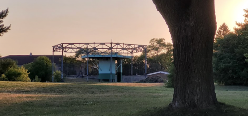 A partially constructed structure in a grassy area, framed by a large tree, with a sunset in the background.