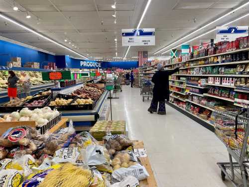 A grocery store aisle filled with produce and various food items, shoppers browsing in a bright, organized space.