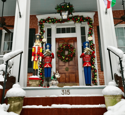 A festive front porch decorated with nutcracker figures, a wreath, and a dog in a sweater, all covered in snow.