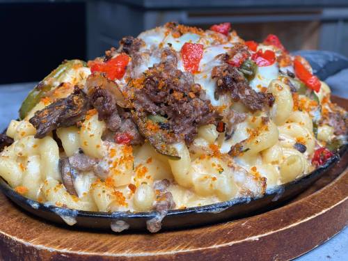 A sizzling skillet of macaroni and cheese topped with seasoned beef, peppers, and melted cheese, garnished with breadcrumbs.