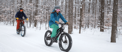 Two people riding fat bikes on a snowy trail through a forest, wearing winter gear and helmets.