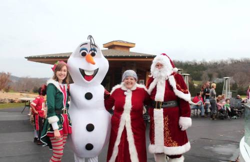 A festive scene with an elf, a snowman, and two people in Santa costumes, smiling together outdoors.