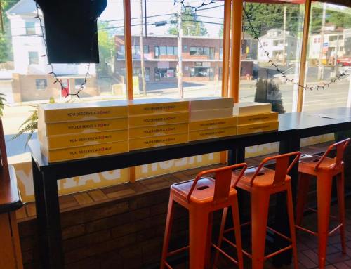 A cozy eatery with orange stools and stacked pizza boxes on a table, featuring a view of the street outside.
