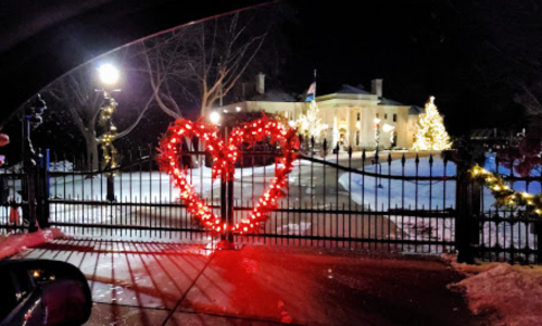 A heart-shaped light display at a gated entrance, with a beautifully lit mansion and trees in the background.