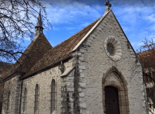A quaint stone church with a steeple, arched windows, and a cross on top, set against a blue sky.