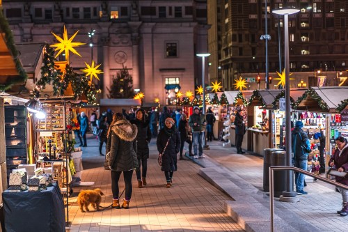 A festive market scene at night, with stalls, holiday decorations, and people walking, including a dog.