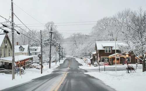 A snowy street scene with quaint houses, power lines, and trees blanketed in white, creating a serene winter atmosphere.