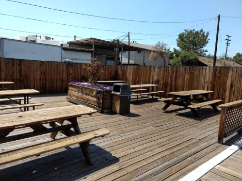 Outdoor seating area with wooden picnic tables, a flower planter, and a trash can, surrounded by a wooden fence.