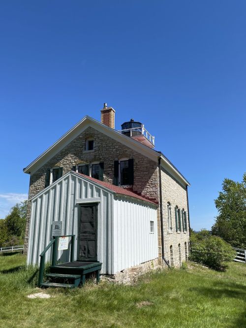 A historic stone house with a white wooden addition, set against a clear blue sky and surrounded by greenery.
