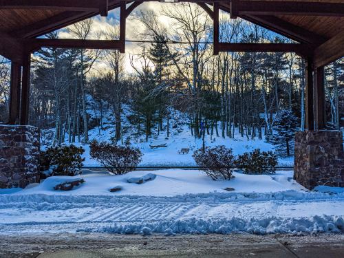 A snowy landscape viewed through a wooden archway, with trees and a bench in the background under a cloudy sky.