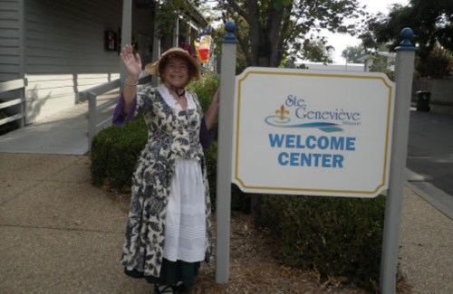 A woman in historical attire stands beside a "Welcome Center" sign in Ste. Genevieve, Missouri.