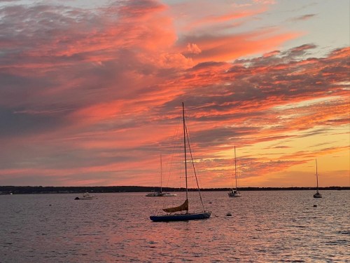 A tranquil sunset over a calm sea, with a sailboat in the foreground and several boats in the distance.