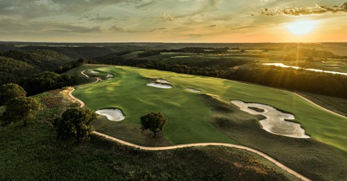 Aerial view of a golf course at sunset, featuring sand traps and lush green fairways surrounded by rolling hills.