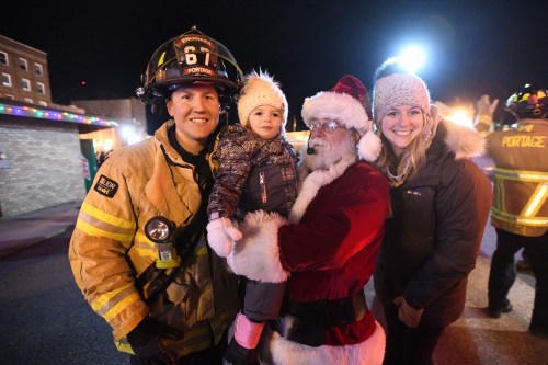 A firefighter, Santa, and a child in winter attire pose together at a festive nighttime event.