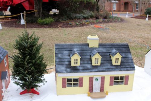 A miniature yellow house with a gray roof and a small Christmas tree beside it, set on a grassy area.