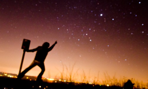 Silhouette of a person pointing at the night sky filled with stars, with city lights in the background.